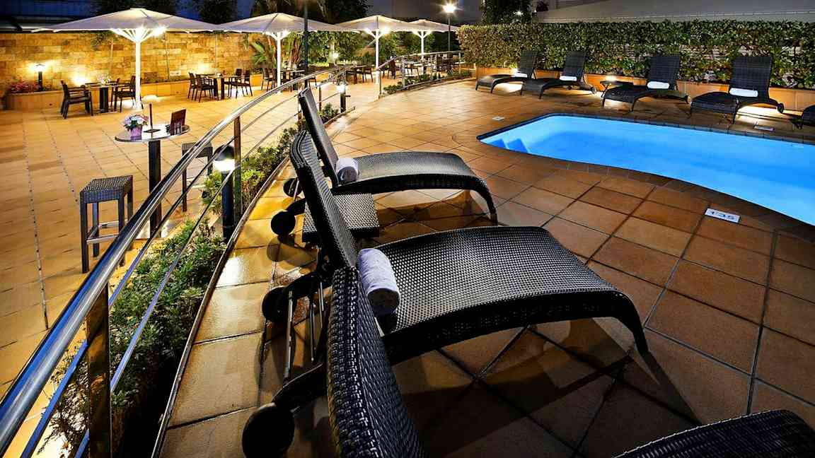 Hotellets poolområde med solstolar och parasoll vid ett upplyst och kaklat uteområde. (AI-genererad text)