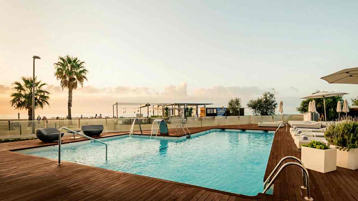 Bilden visar en utomhuspool med trädäck och solsängar vid kanten. I bakgrunden syns ett par palmer, en byggnad och havet, allt under en ljus kvällshimmel. (AI-genererad text)