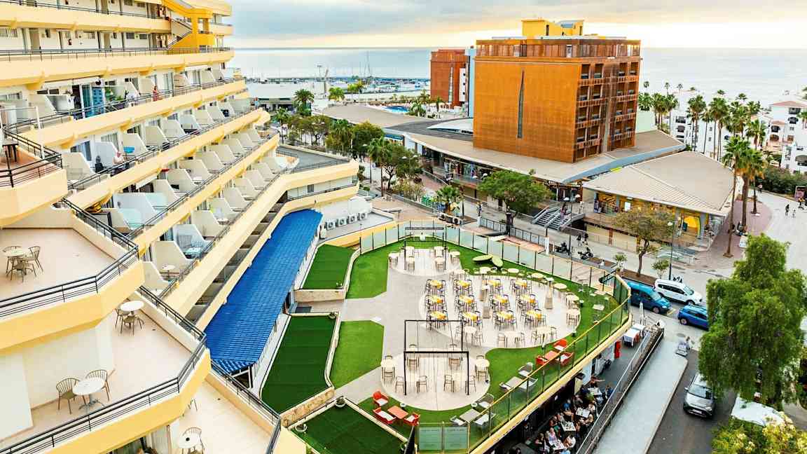 Hotell med gula fasader och balkonger, en terrass med bord och stolar omgiven av grönska, och en vy över staden och havet i bakgrunden. (AI-genererad text)