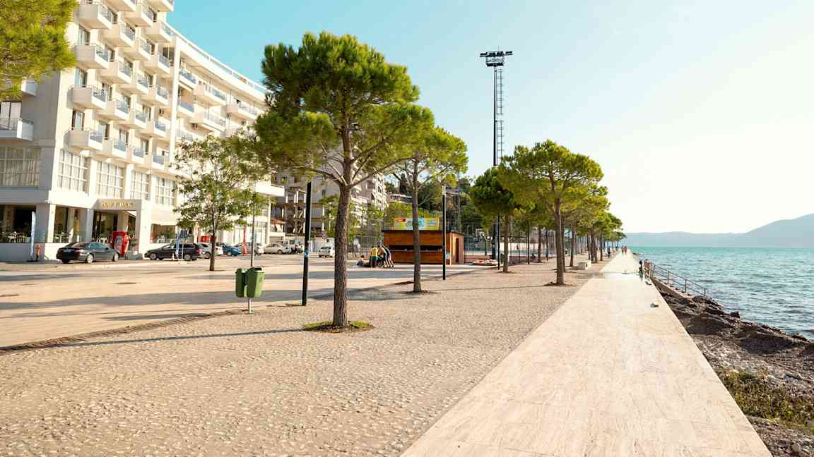 En strandpromenad med träd och vitkaklat golv längs vattnet, med en modern byggnad till vänster. (AI-genererad text)