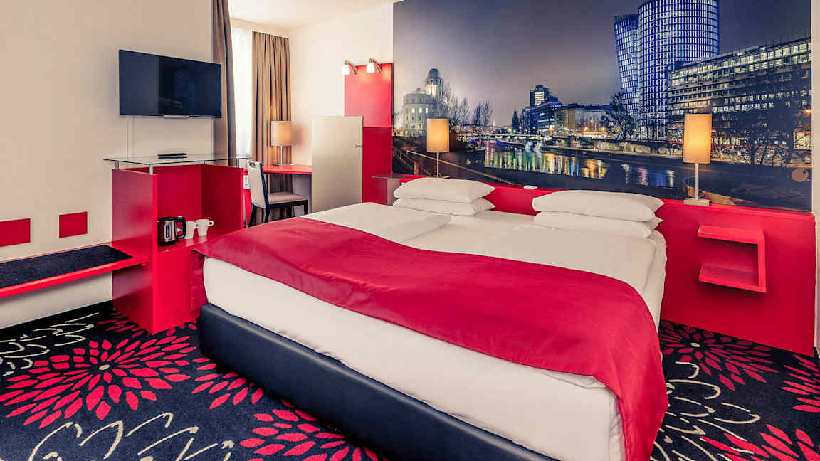 Ett hotellrum med en dubbelsäng, en TV på väggen och ett skrivbord bredvid fönstret. Väggarna är dekorerade med en storbild av en stadslandskap. (AI-genererad text)