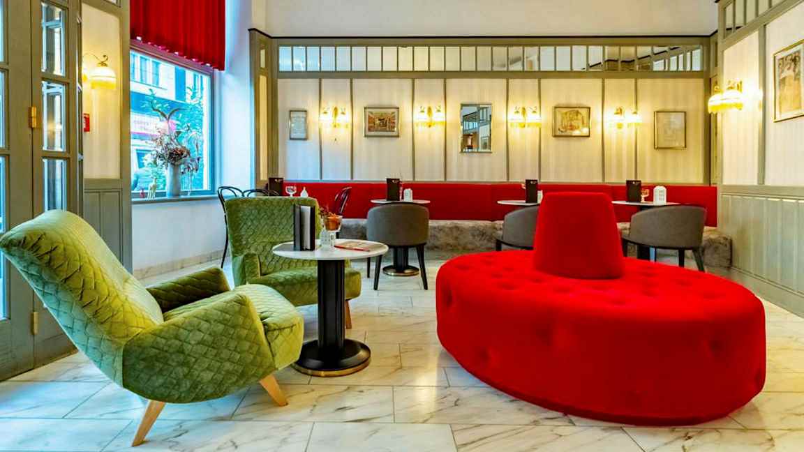 En hotellobby med sittgrupp inkluderande gröna fåtöljer, röda och gråa sittmöbler och marmorgolv. (AI-genererad text)