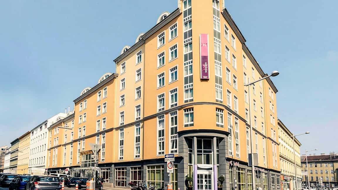 En ljusgul flerfaldigt fönstrad byggnad med skylt och rundad hörningsfasad. (AI-genererad text)