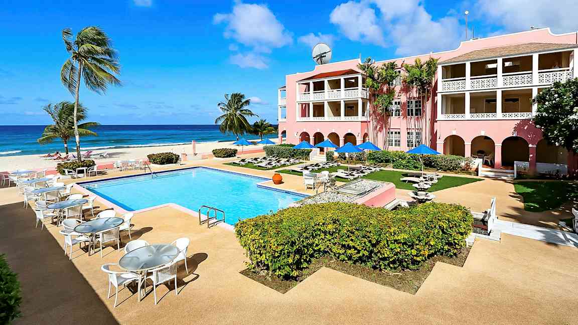 Bild av en solig resort med en utomhuspool, omgivande uteplatser och ett rosa hotell med balkonger, beläget vid havet. (AI-genererad text)