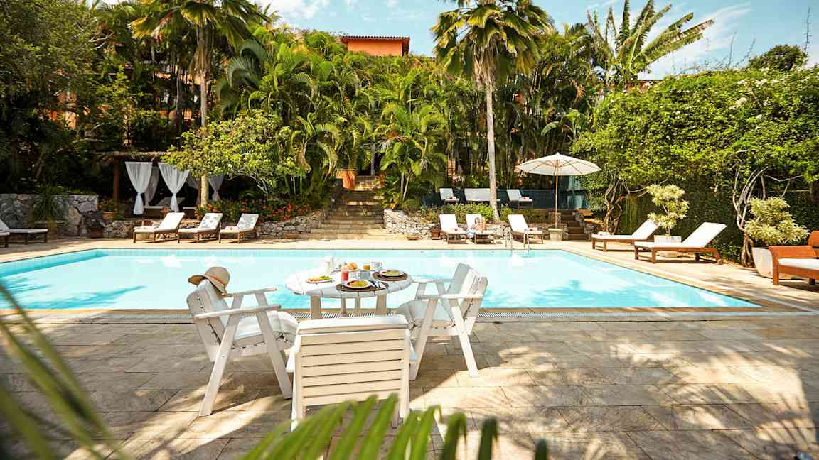 En utomhuspool omgiven av grönska och solstolar. Ett bord med mat och dryck står vid poolkanten. (AI-genererad text)