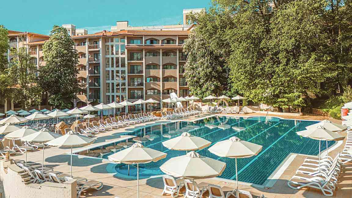 Poolområde med solstolar och parasoller, bakom syns en hotellbyggnad och gröna träd. (AI-genererad text)