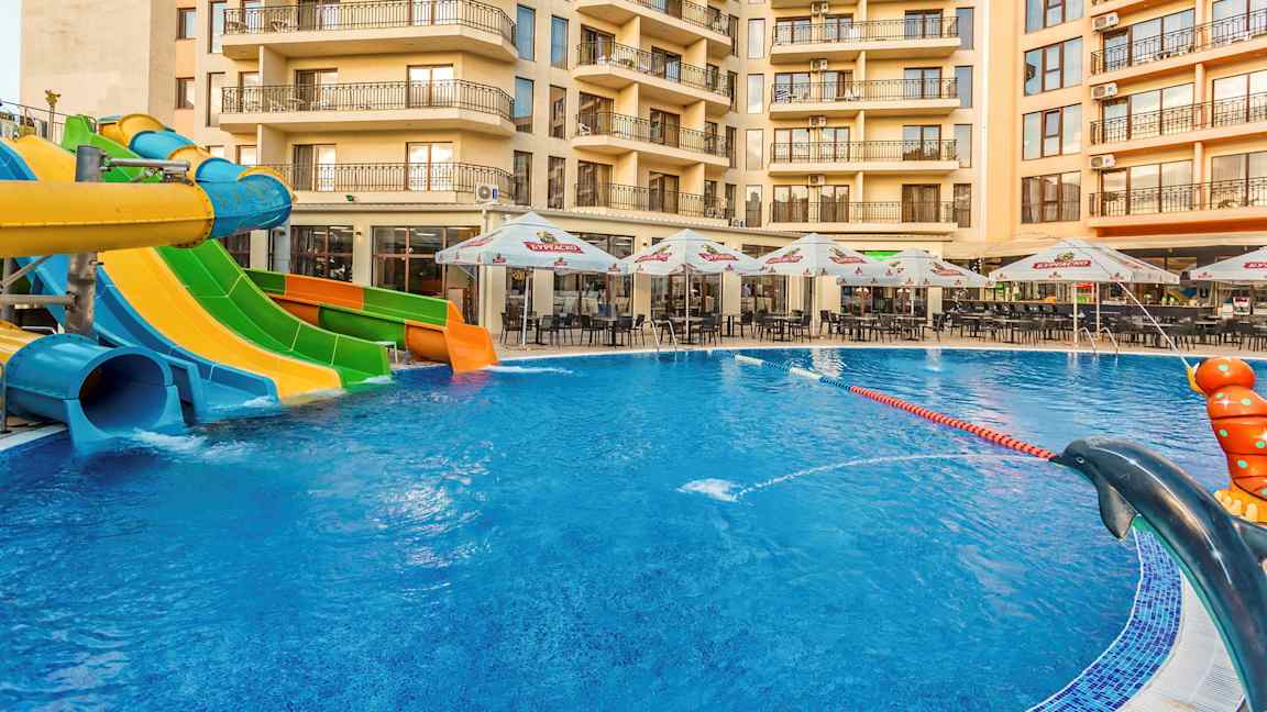 Bilden visar en swimmingpool med vattenrutschbanor i olika färger framför ett hotell, med utemöbler och parasoller vid poolområdet. (AI-genererad text)