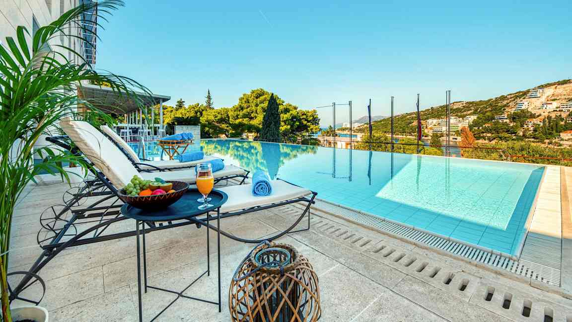 En utsikt mot en infinitypool med solstolar, bord med frukt och dryck intill sig, samt gröna omgivningar och himmel i bakgrunden. (AI-genererad text)