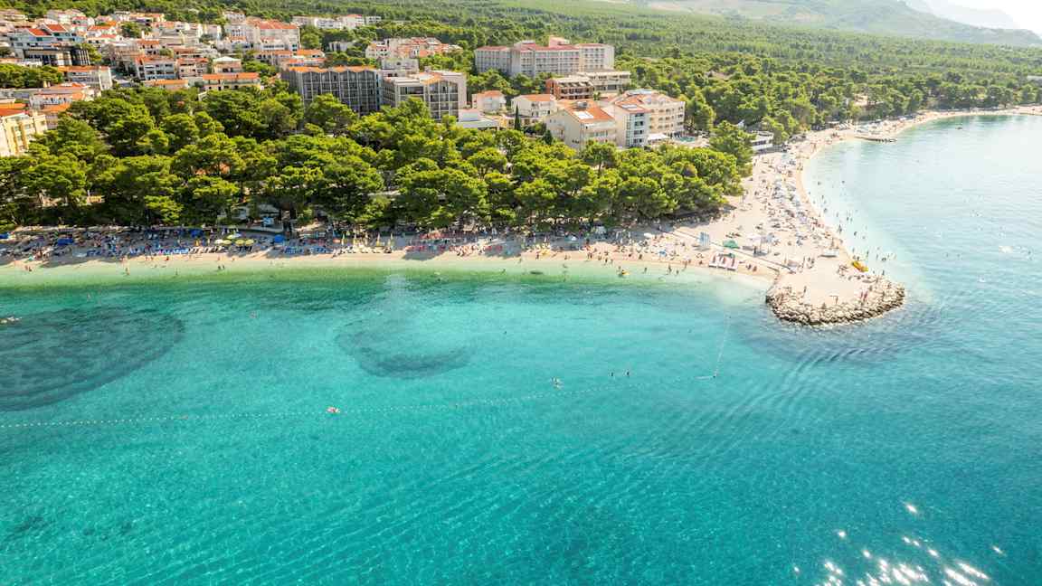 Luftbild av en kust trots klarblått vatten, strand med solstolar och en tät bebyggelse omgiven av skog och berg. (AI-genererad text)