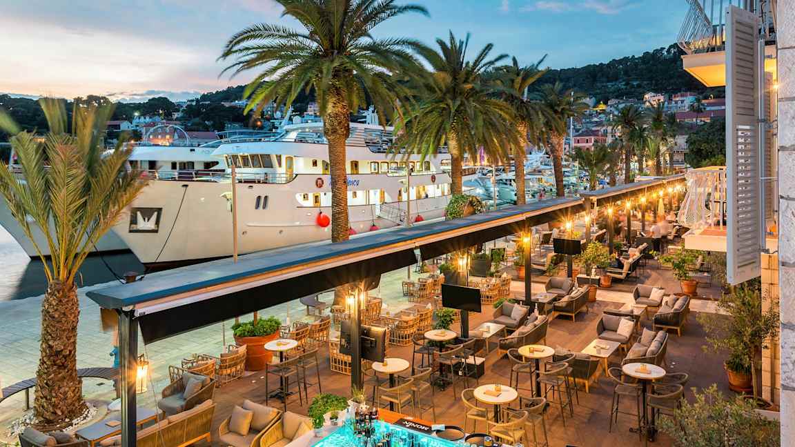 En vy av en utomhusrestaurang vid en hamn med palmer och stora båtar. (AI-genererad text)