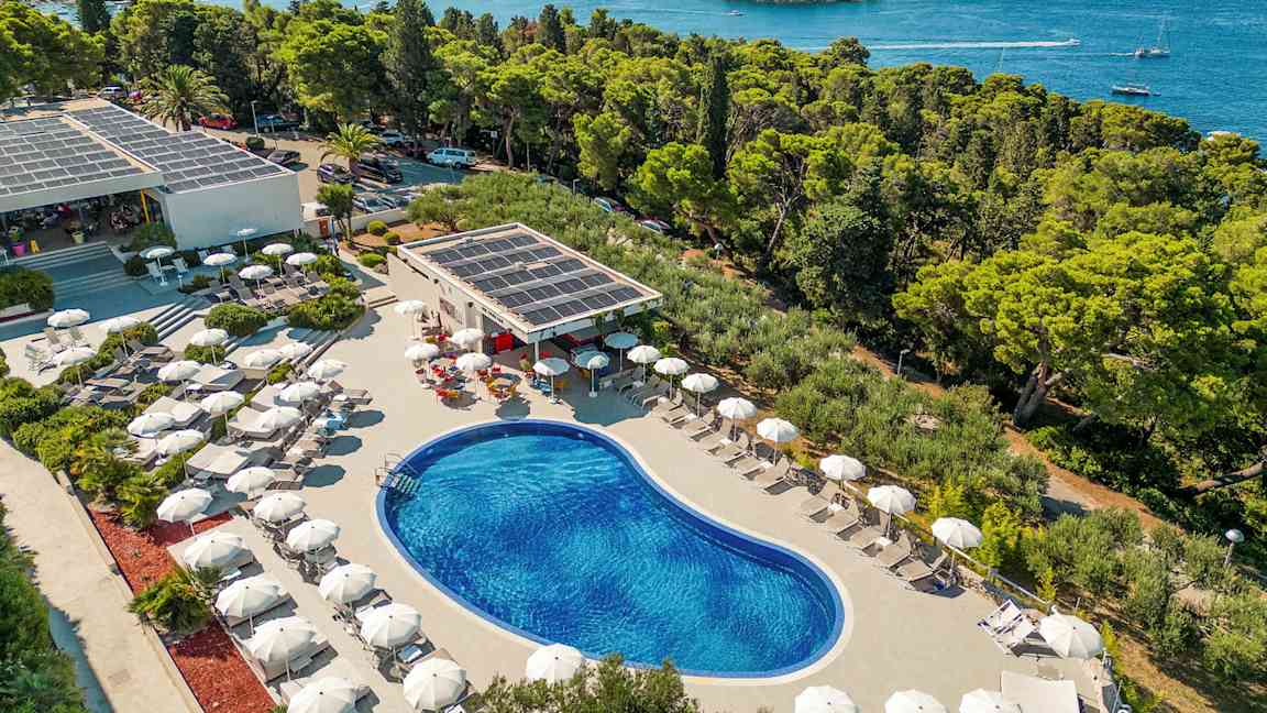 Vy över en utomhuspool omgiven av solstolar med parasoll, belägen nära kusten med utsikt över skog och hav. (AI-genererad text)