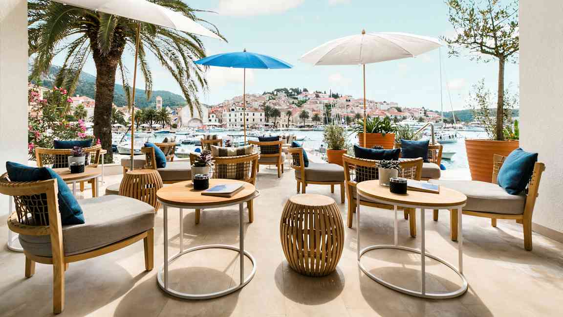 Uteservering med bord och stolar under parasoller, omgiven av palmer med utsikt över en pittoresk hamn och stad. (AI-genererad text)