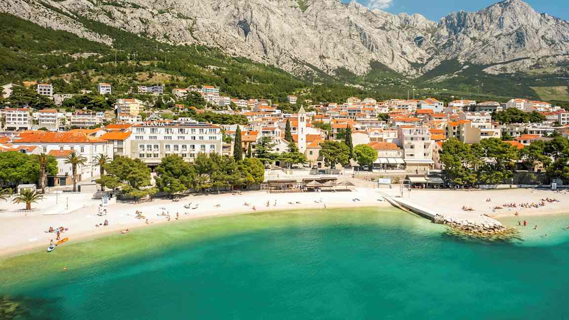 En kuststad vid foten av ett berg med en sandstrand och klart turkost vatten. (AI-genererad text)