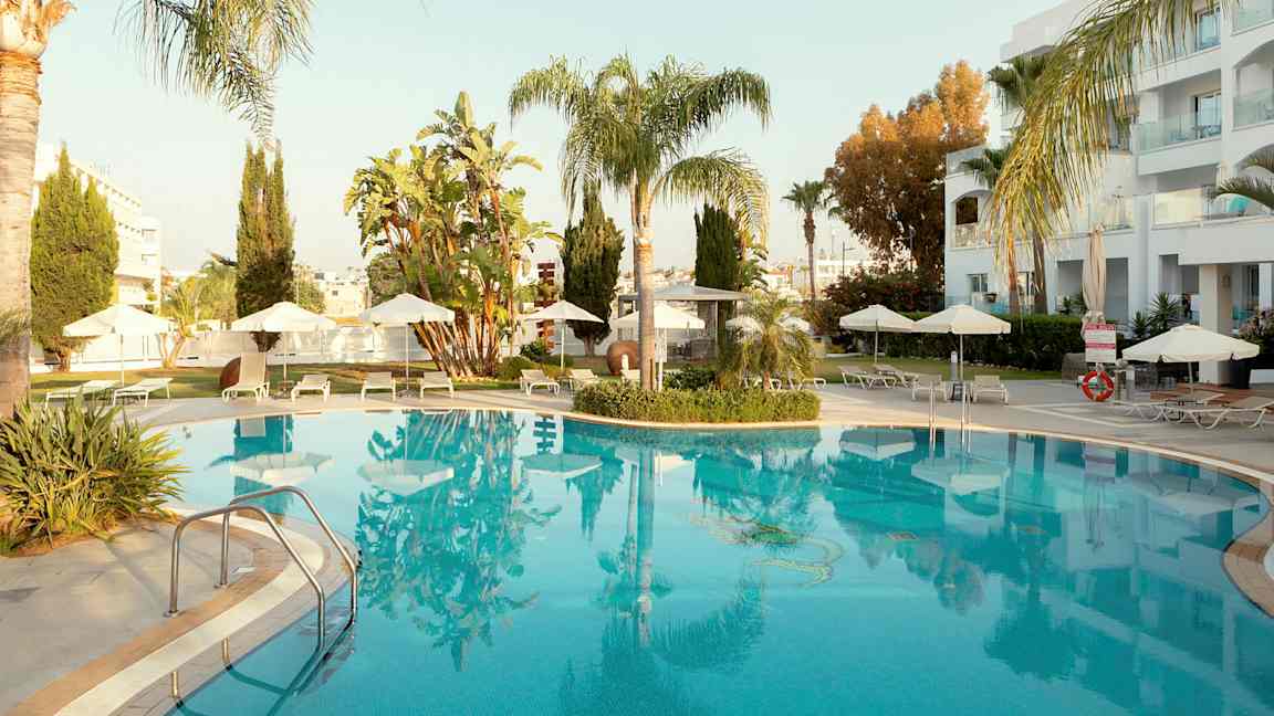 Utomhuspool omgiven av solstolar och parasoller, med palmer och byggnader i bakgrunden. (AI-genererad text)