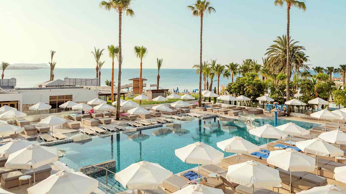 Utomhuspoolområde med solstolar och parasoller, omgiven av palmer och nära havet. (AI-genererad text)