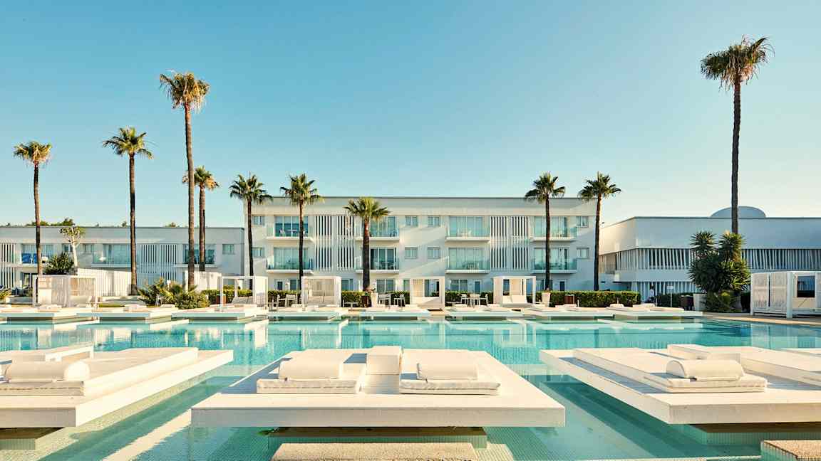 En modern byggnad med balkonger, omgiven av palmer och en swimmingpool med solstolar. (AI-genererad text)