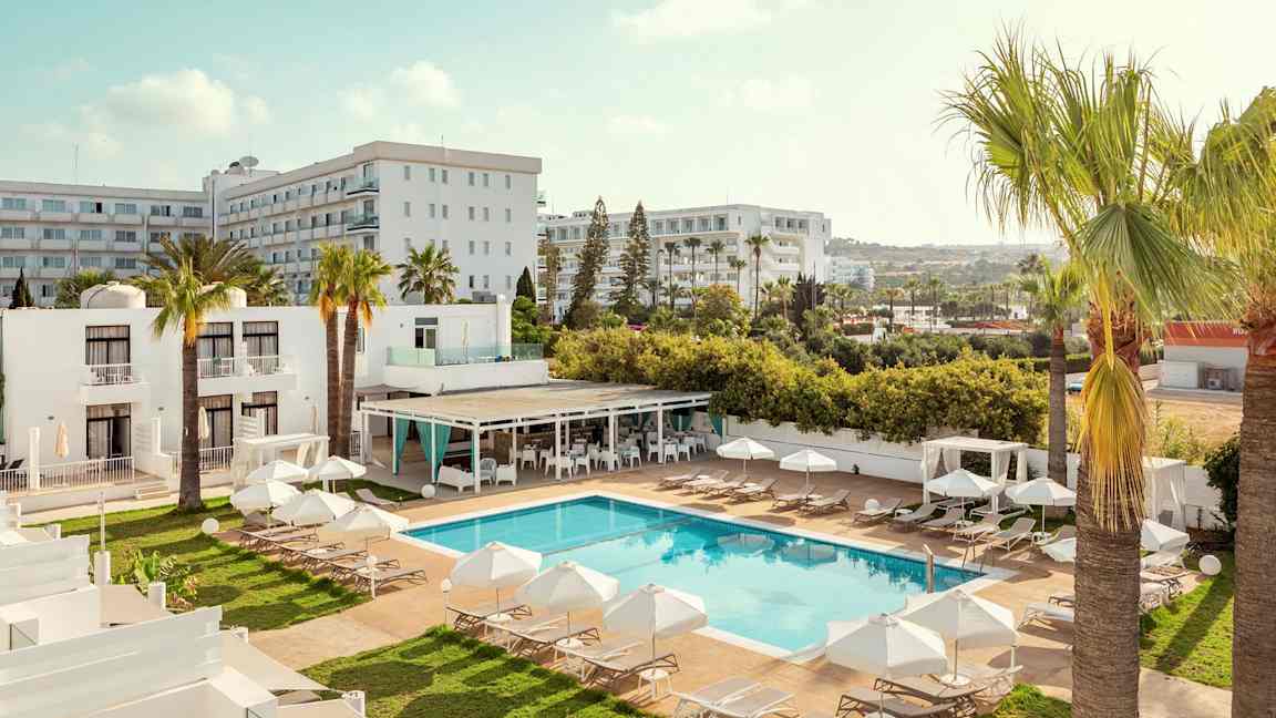 Ett hotellområde med pool, solstolar och parasoller, omgiven av palmer och byggnader. (AI-genererad text)