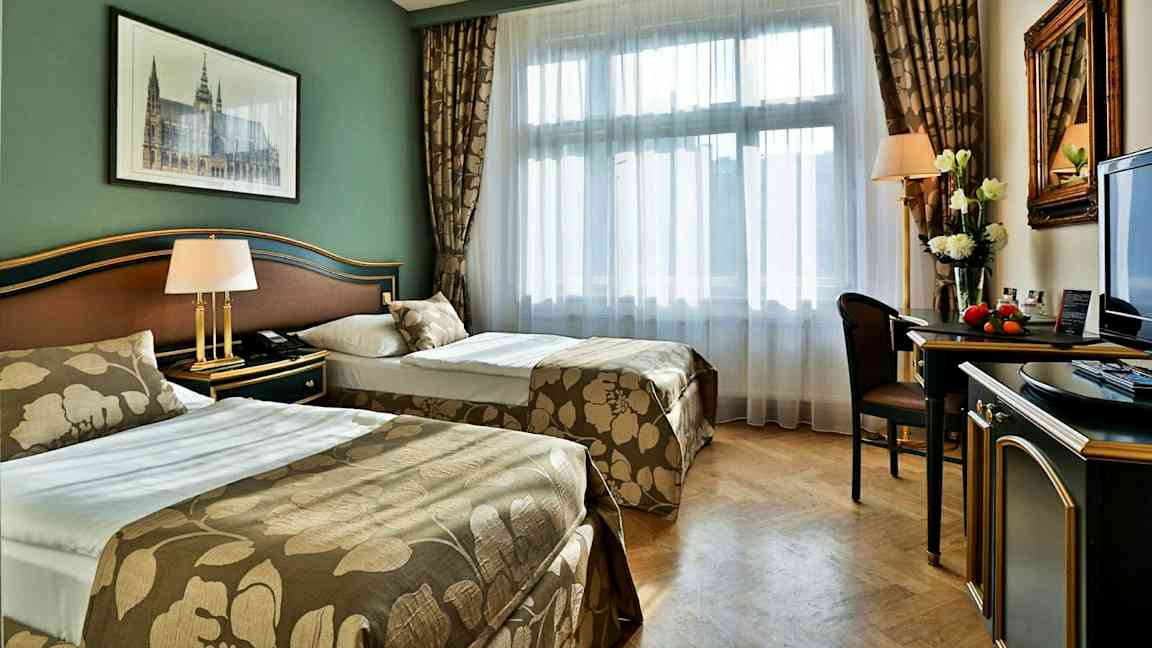 Ett hotellrum med två separata sängar, grön vägg, dekorativt sängtäcke, skrivbord och fönster med ljus insläpp. (AI-genererad text)
