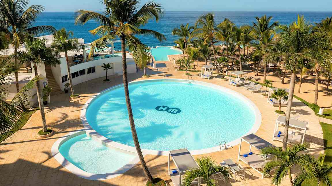 Ett hotellområde med en stor rund pool omgiven av palmer, solsängar och cabanas. Utsikt över havet i bakgrunden. (AI-genererad text)