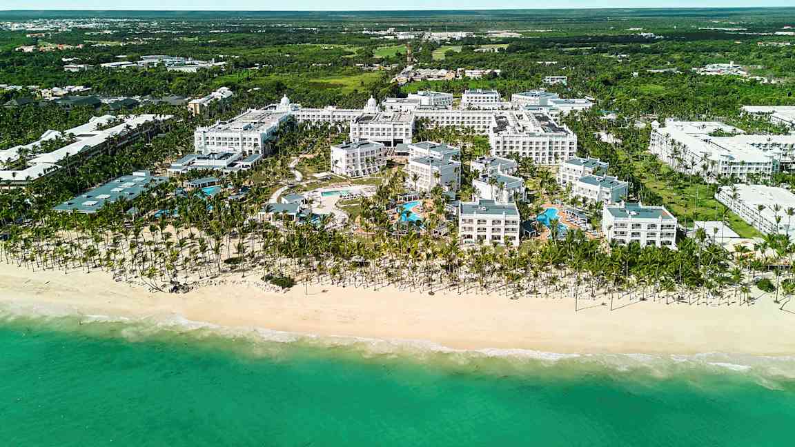 Flygbild av ett hotellkomplex vid en strand inklusive poolområde, omgiven av grönska. (AI-genererad text)