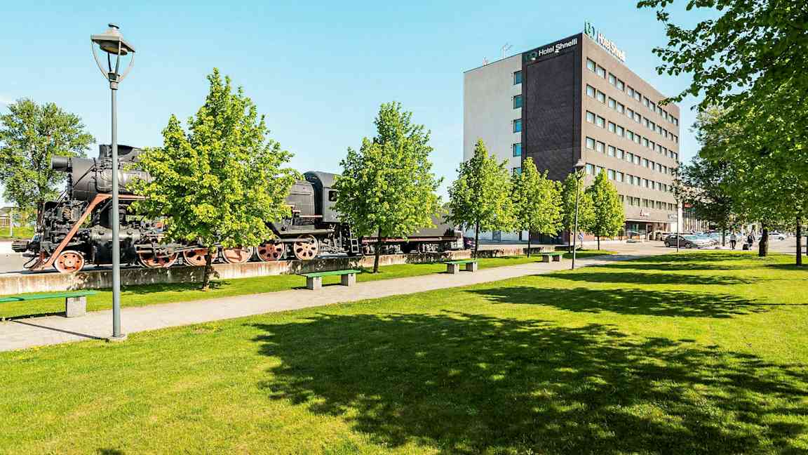 Ett gammalt ånglok står utomhus bredvid en modern hotellanläggning och grönskande träd. (AI-genererad text)