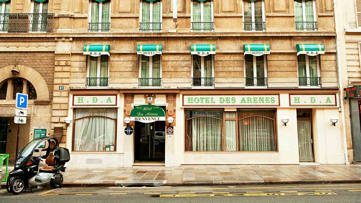 Byggnad med hotellfasad, skyltar med texten 'Hotel des Arenes' och grönvita markiser ovanför fönstren. (AI-genererad text)