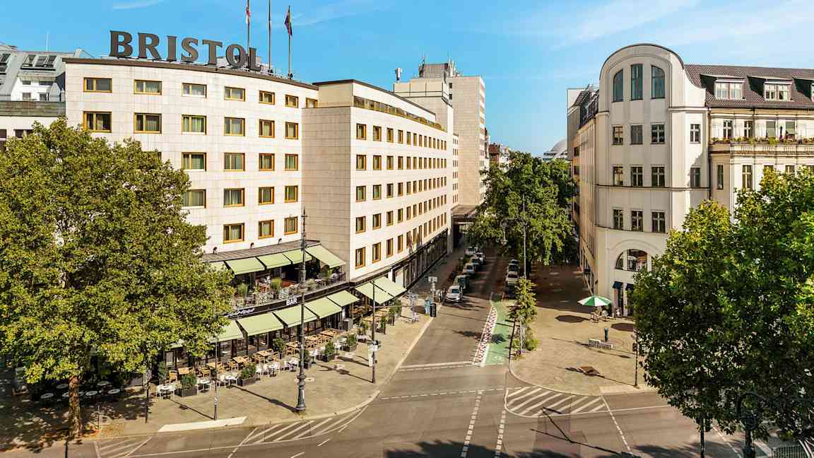 Ett stadslandskap som visar en byggnad med texten "Bristol" på taket, omgiven av träd och en gata med uteservering. (AI-genererad text)