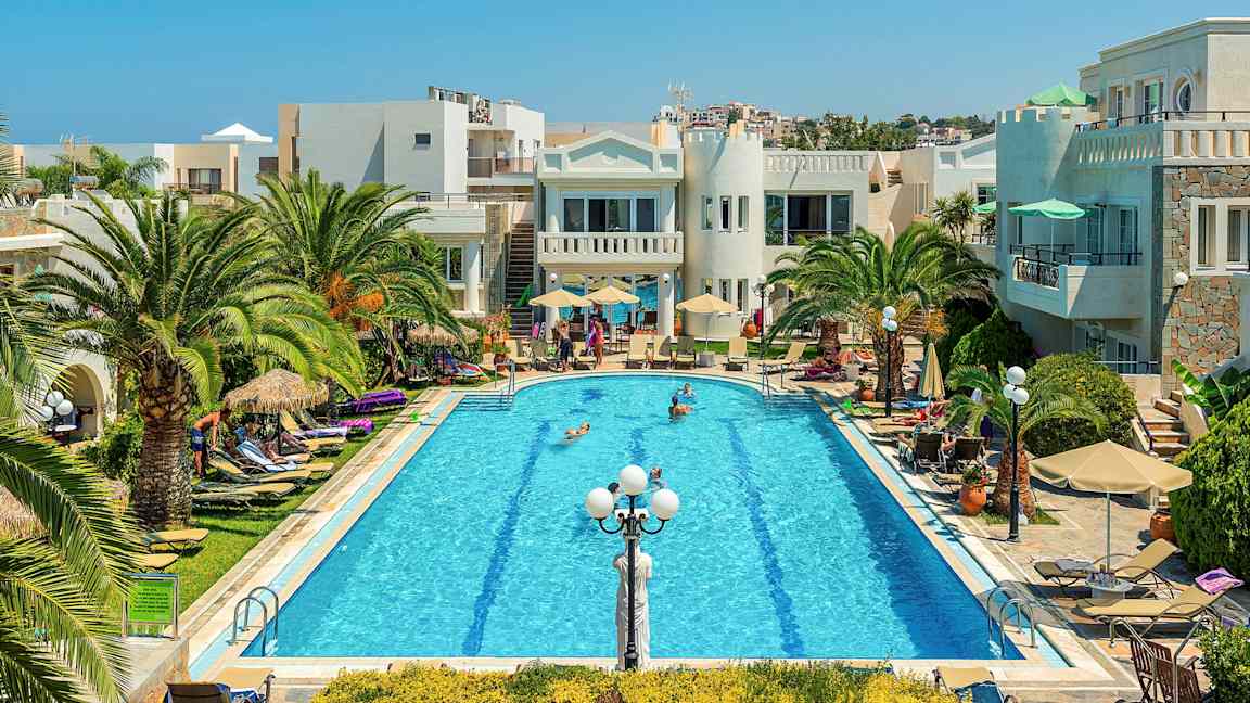 En pool omgiven av palmer, solstolar och byggnader under en klarblå himmel. (AI-genererad text)