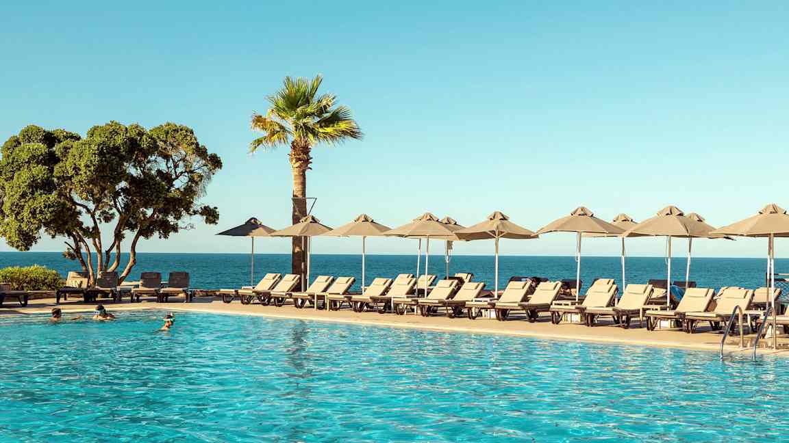 En pool i förgrunden med solstolar och parasoller vid sidan. Bakom syns havet och en klar blå himmel. (AI-genererad text)