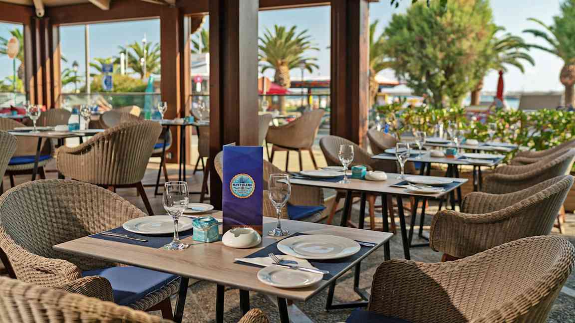 En uterestaurang med dukade bord, rottingstolar och utsikt över träd och palmer. (AI-genererad text)