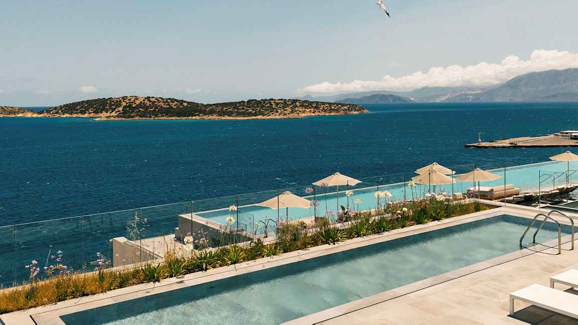 Bild av en infinitypool med utsikt över havet och omgivande öar. En glasbalustrad omger poolområdet, och parasoller finns vid poolkanten. (AI-genererad text)