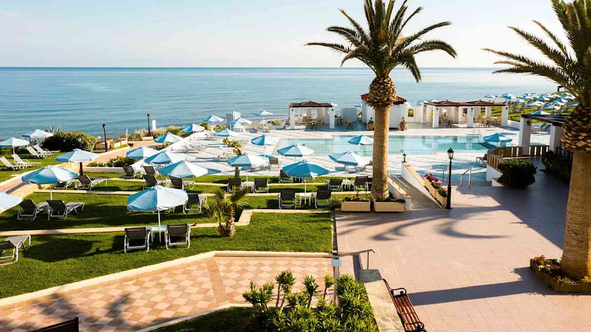 Ett hotellområde med en pool och solstolar under blå parasoller, omgärdad av palmer och med utsikt över havet. (AI-genererad text)