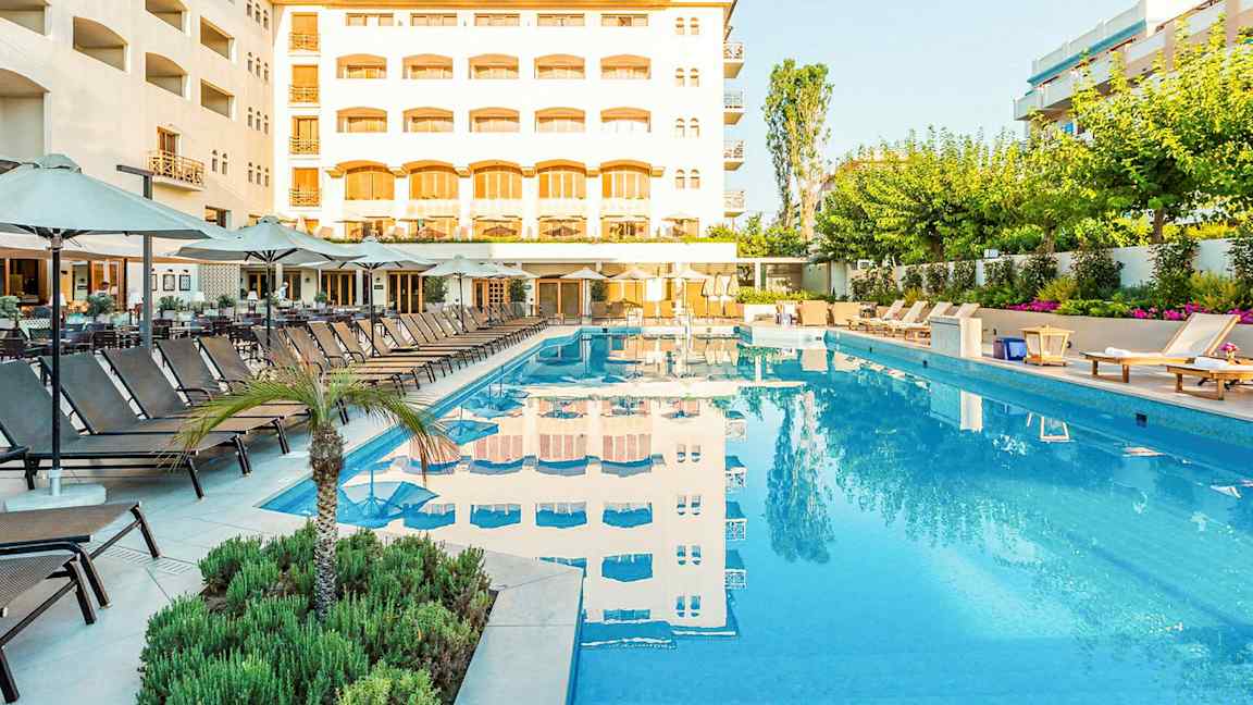 En utomhuspool med omgivande solstolar och parasoller framför ett hotell. (AI-genererad text)