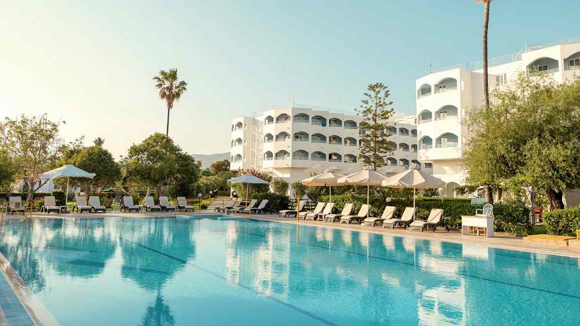 Hotellbyggnad med pool, solstolar och parasoller i förgrunden. (AI-genererad text)