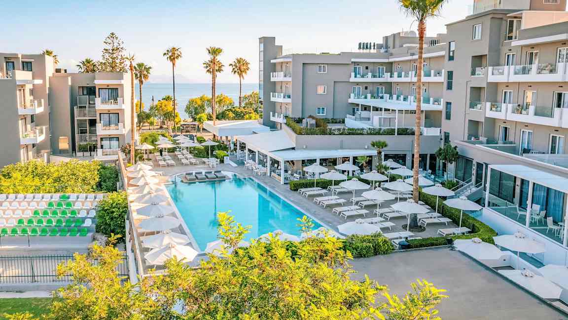 Ett modernt hotellkomplex med pool, solstolar och parasoller, omgiven av grönska och med utsikt över havet. (AI-genererad text)