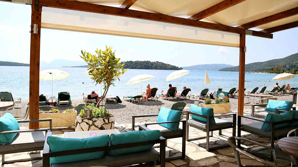 Utsikt från ett skuggigt sittområde mot en strand med solstolar och parasoller vid en lugn havsvik. (AI-genererad text)