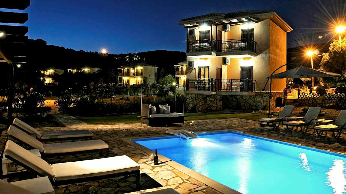 En kvällsbild av en utomhuspool och flera solstolar. Bakgrunden visar ett hus med balkonger och omgivande belysning. (AI-genererad text)