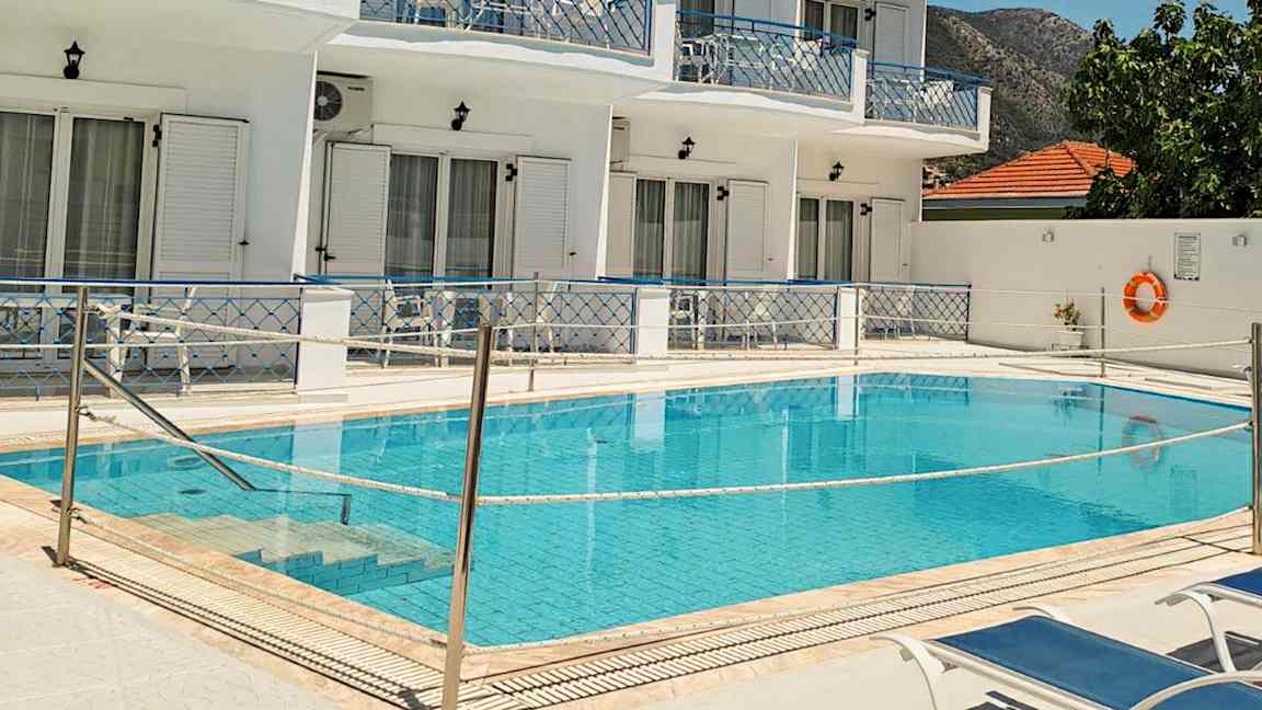 Bild av en pool framför ett vitmålat hotell med blåa balkonger och solstolar vid poolen. (AI-genererad text)
