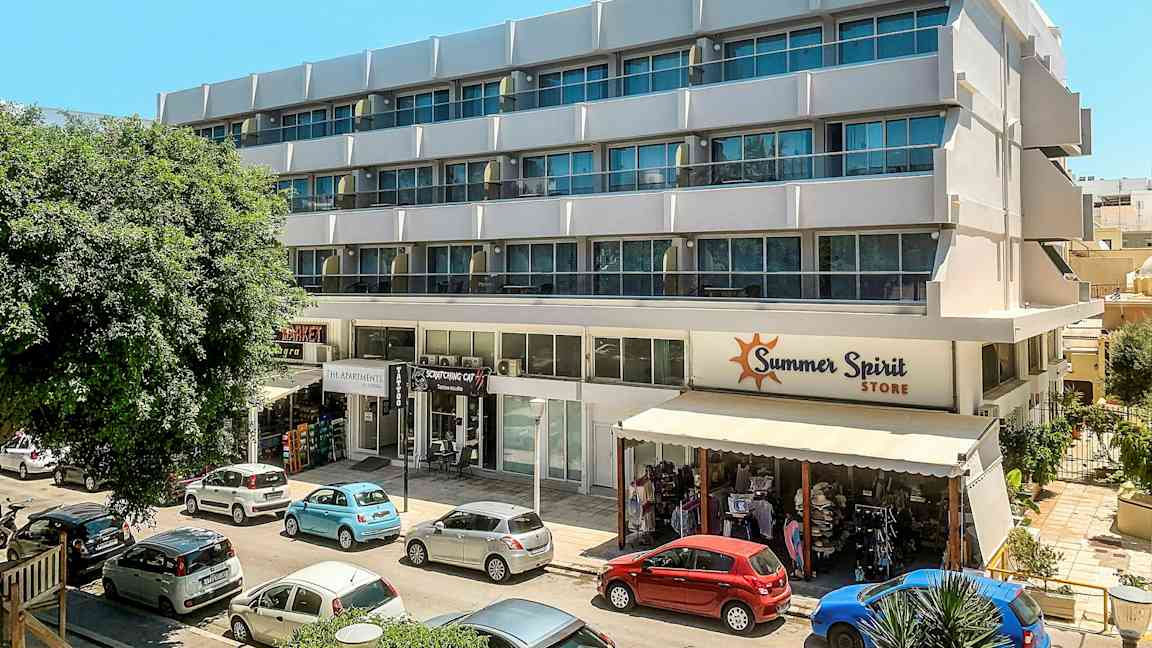 En modern byggnad med flera våningar och affärer längs bottenvåningen. Framför byggnaden står parkerade bilar. (AI-genererad text)