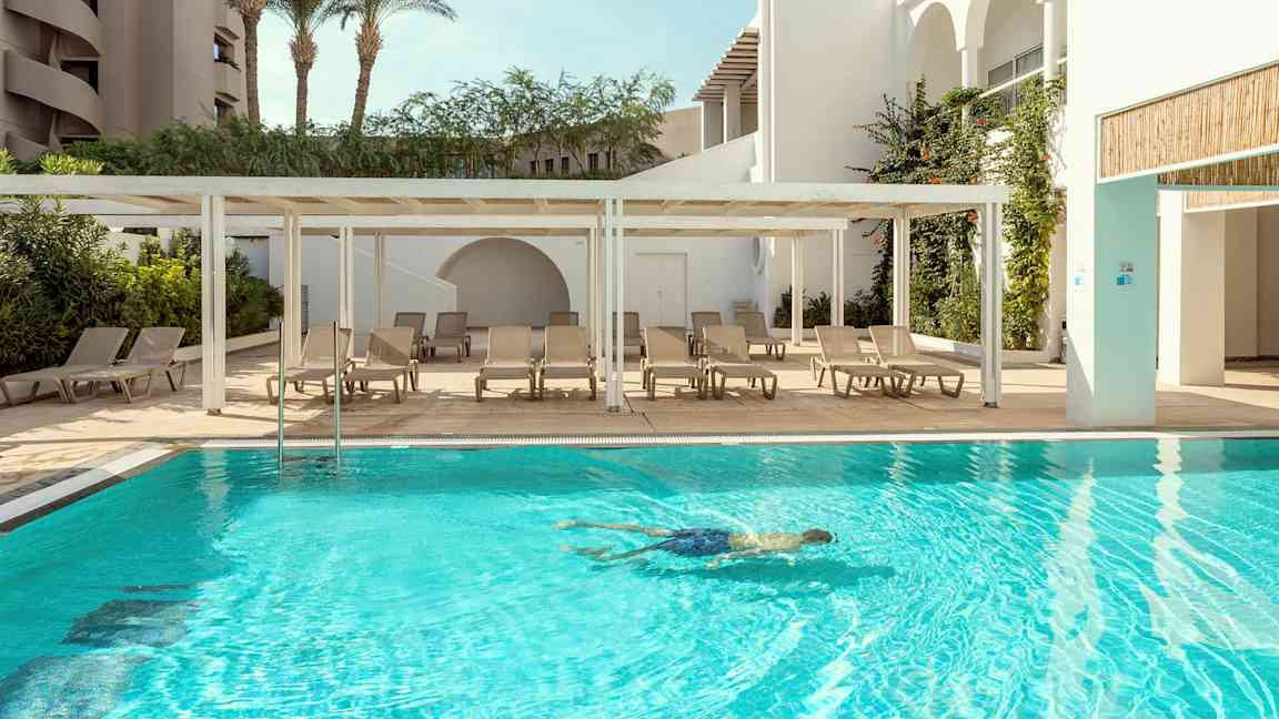 En utomhuspool med solstolar bredvid och en person som simmar i vattnet. (AI-genererad text)