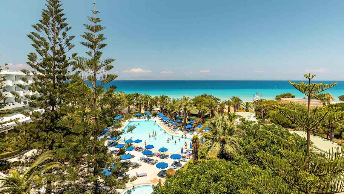 Bild av hotellområde med pool och solsängar omgivna av grönska och utsikt mot havet. (AI-genererad text)