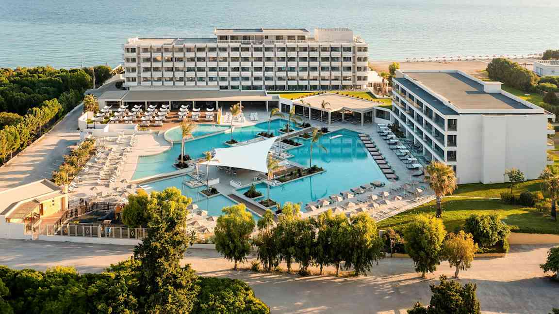 Flygfoto av ett hotellkomplex med poolområde och hav i bakgrunden. (AI-genererad text)