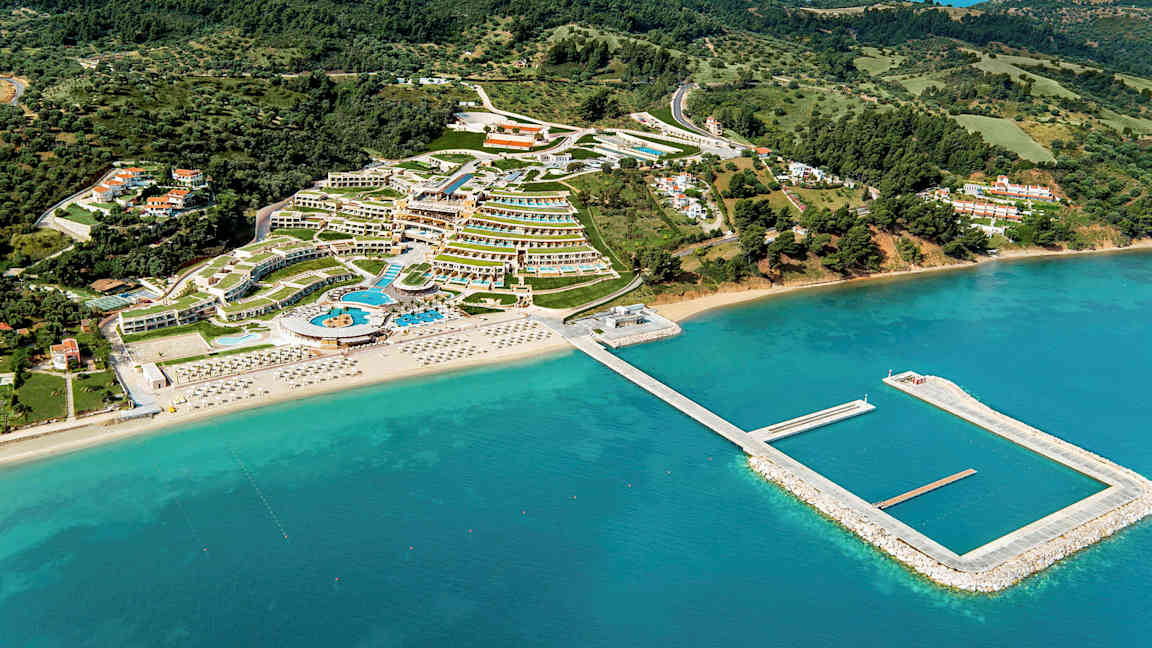 Flygbild över ett semesterområde med hotell, pooler och strand med solstolar, omgivet av grönska och hav. (AI-genererad text)