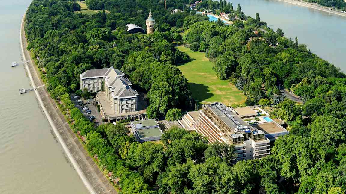 Luftbild av en grönskande ö med byggnader, gräsytor och träd omgiven av en flod. (AI-genererad text)