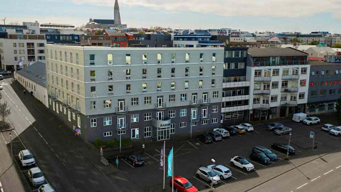 Flera moderna byggnader omger ett öppet parkeringsområde, med bilar parkerade på marken och en kyrka synlig i bakgrunden. (AI-genererad text)