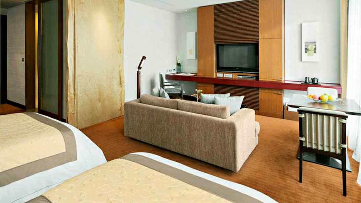 Ett hotellrum med två sängar, soffa, ett skrivbord och en TV på en träpanel. (AI-genererad text)