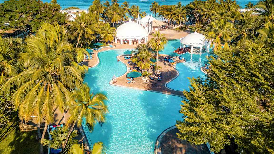 En stor pool med flera sektioner omgivet av palmer och byggnader med vita kupoler. I bakgrunden syns havet. (AI-genererad text)
