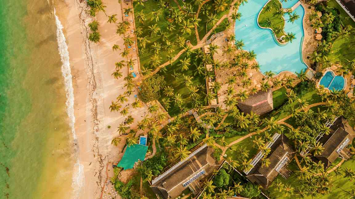 Flygbild av en resort vid stranden med gröna gräsmattor, palmer och en stor pool. (AI-genererad text)