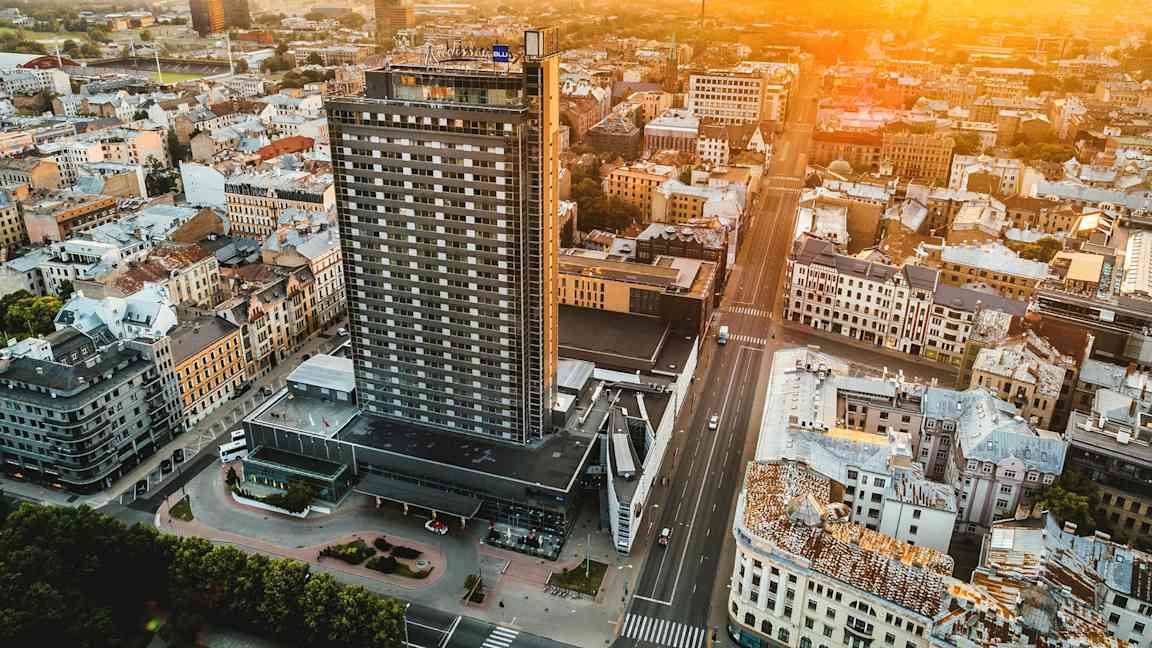 Flygfoto av en stad med en hög byggnad i framgrunden och andra byggnader omkring, samtidigt som solen går ner vid horisonten. (AI-genererad text)