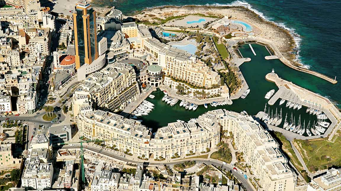 Flygfoto över en kuststad med höga byggnader, småbåtshamn och havet i bakgrunden. (AI-genererad text)